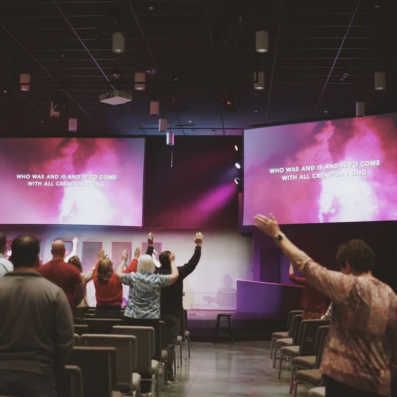 A congregation stands between rows of chairs in a modern church, their hands raised in worship. Two large screens show song lyrics under purple stage lights.